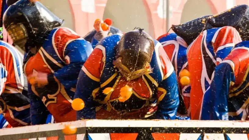 Participantes da Batalha das Laranjas em Ivrea durante o Carnaval na Itália, vestindo capacetes e túnicas de proteção enquanto lançam laranjas uns nos outros em praça pública.