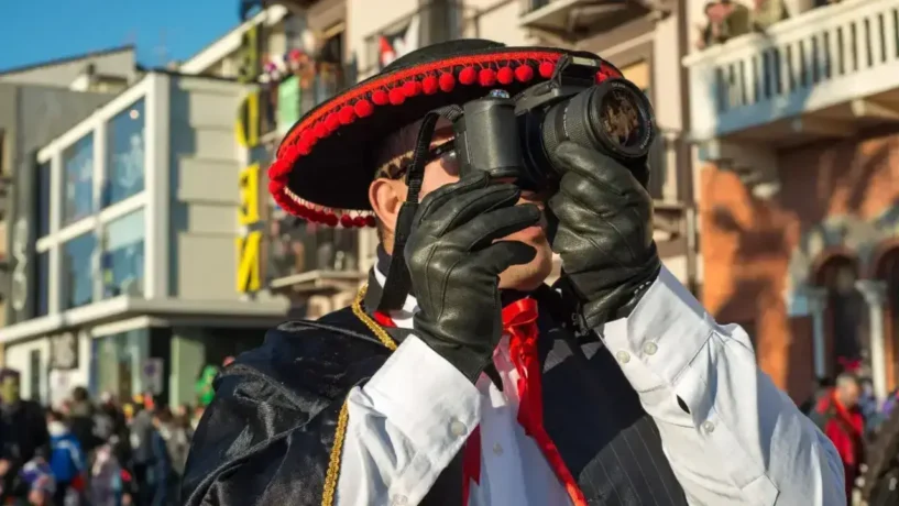 Figurante vestindo fantasia satírica com sombrero preto e pompons vermelhos durante o Carnaval de Viareggio na Itália, fotografando os carros alegóricos de papier-mâché entre a multidão.