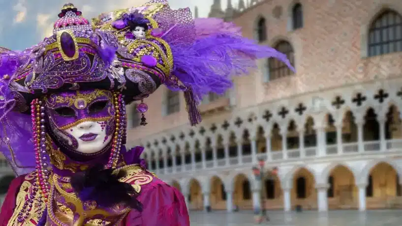 Pessoa vestindo fantasia elaborada de Carnaval na Itália com máscara veneziana ornada em roxo e dourado e penas, posando em frente ao Palácio Ducal na Praça de São Marcos em Veneza.