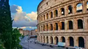Imagem de uma vista do Coliseu em Roma, com céu azul e nuvens, destacando a história e beleza da cid.
