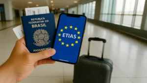 Mão segurando um passaporte brasileiro e um smartphone com o logotipo do ETIAS na tela, em um aeroporto com uma mala de viagem ao fundo.