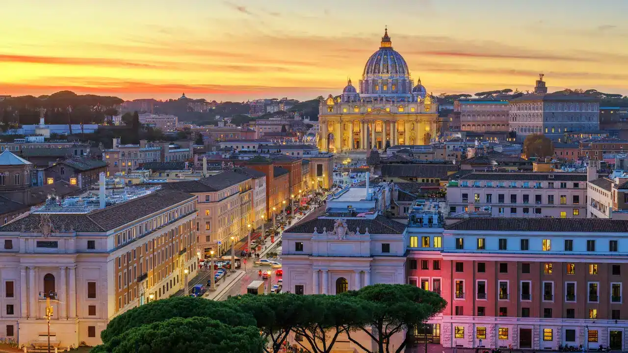 Imagem do Vaticano em Roma ao entardecer, com a cúpula de São Pedro iluminada ao fundo.